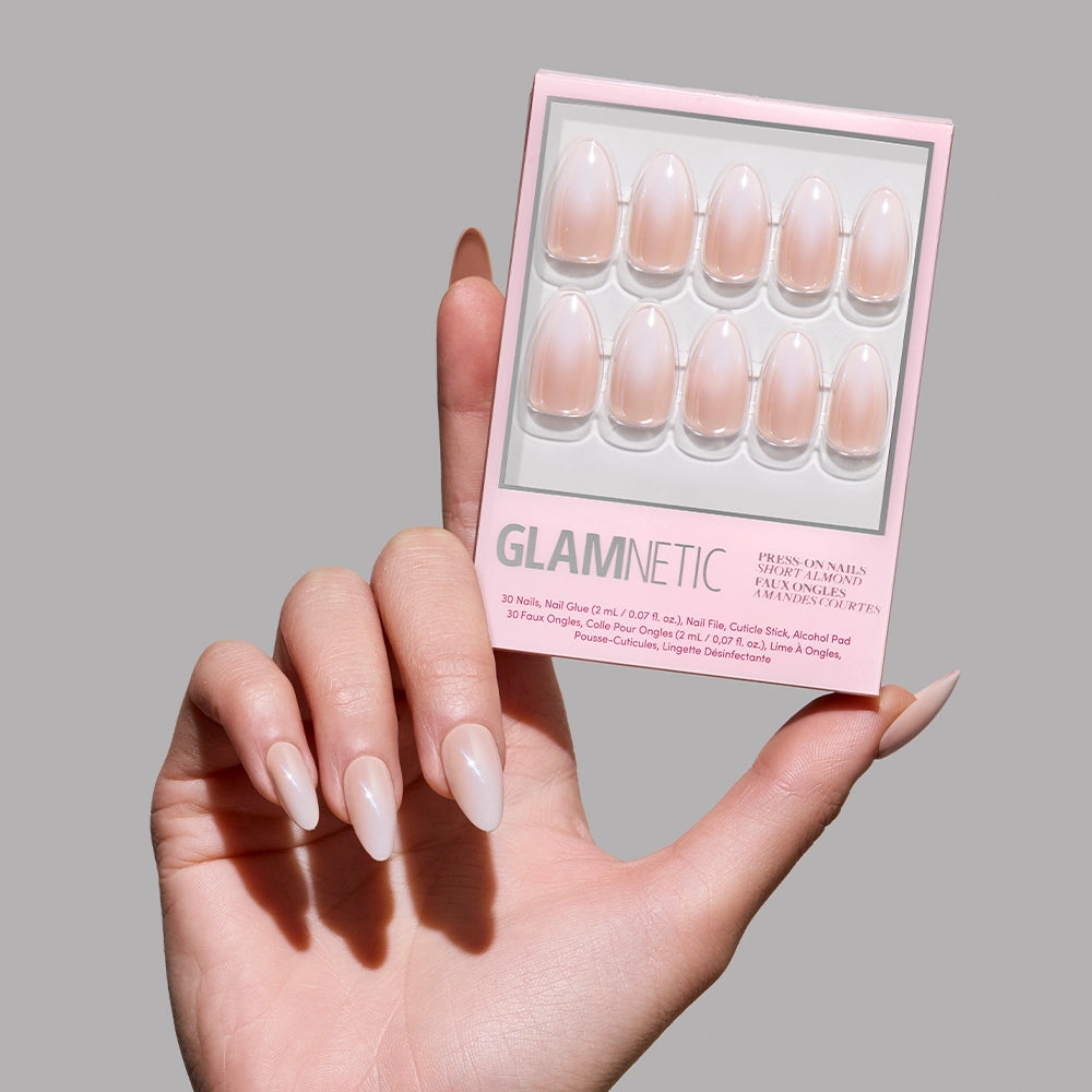 A hand with long, glossy, almond-shaped nude glamnetic Fiancéd press-on nails holds the box against a light pink background, displaying elegant French tips for a classic look.